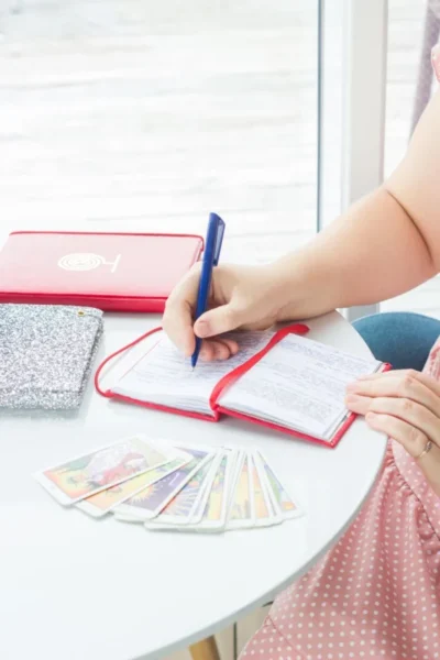 Tarot Journaling: A Guide to Connecting with Your Tarot Deck - 4 A woman writes in a journal with tarot cards displayed on a table. Bright daylight setting.